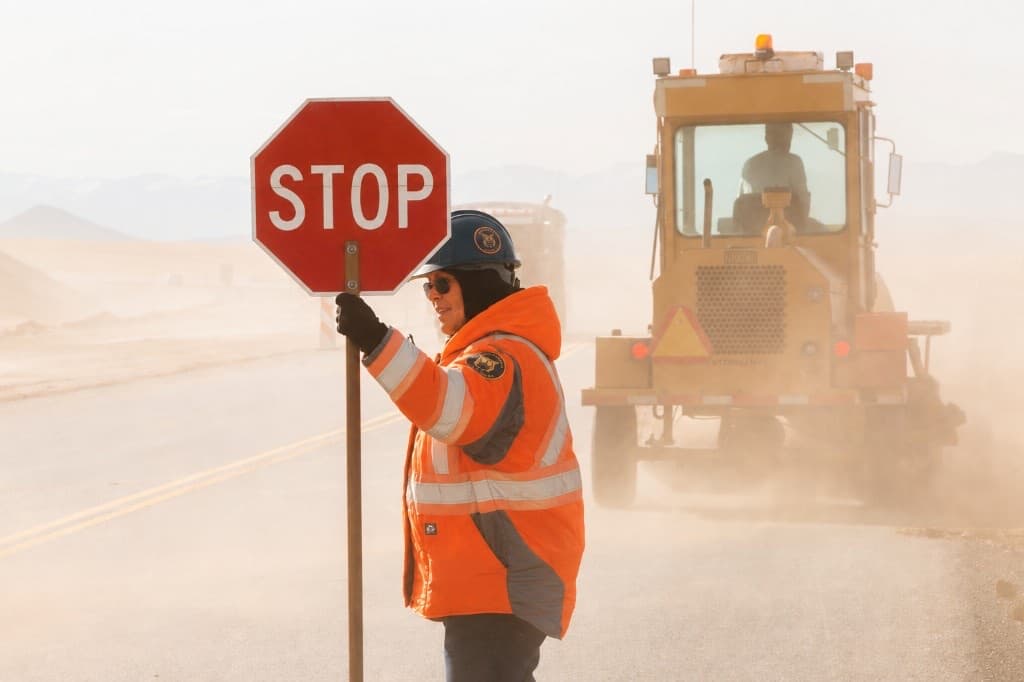 Waylert work-zone flagger in an orange safety jacket holding a STOP sign beside road construction equipment